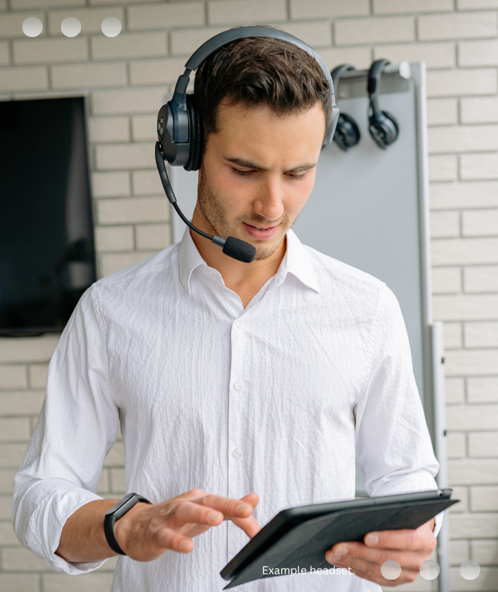 A professional in a white shirt wearing a headset, looking down at a tablet computer while standing in front of a white brick wall