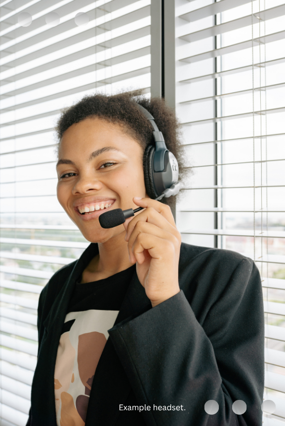 A smiling professional wearing a communication headset with a microphone, standing in front of office window blinds.