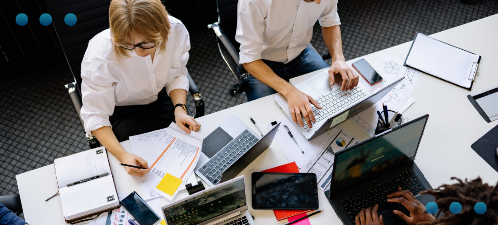 A high-angle, overhead view of a team collaborating at a large, white office desk. The workspace is cluttered with multiple laptops, tablets, notebooks, documents, and charts. Three people are visible, actively working, writing on papers, and typing on their computers in a busy, professional office setting.