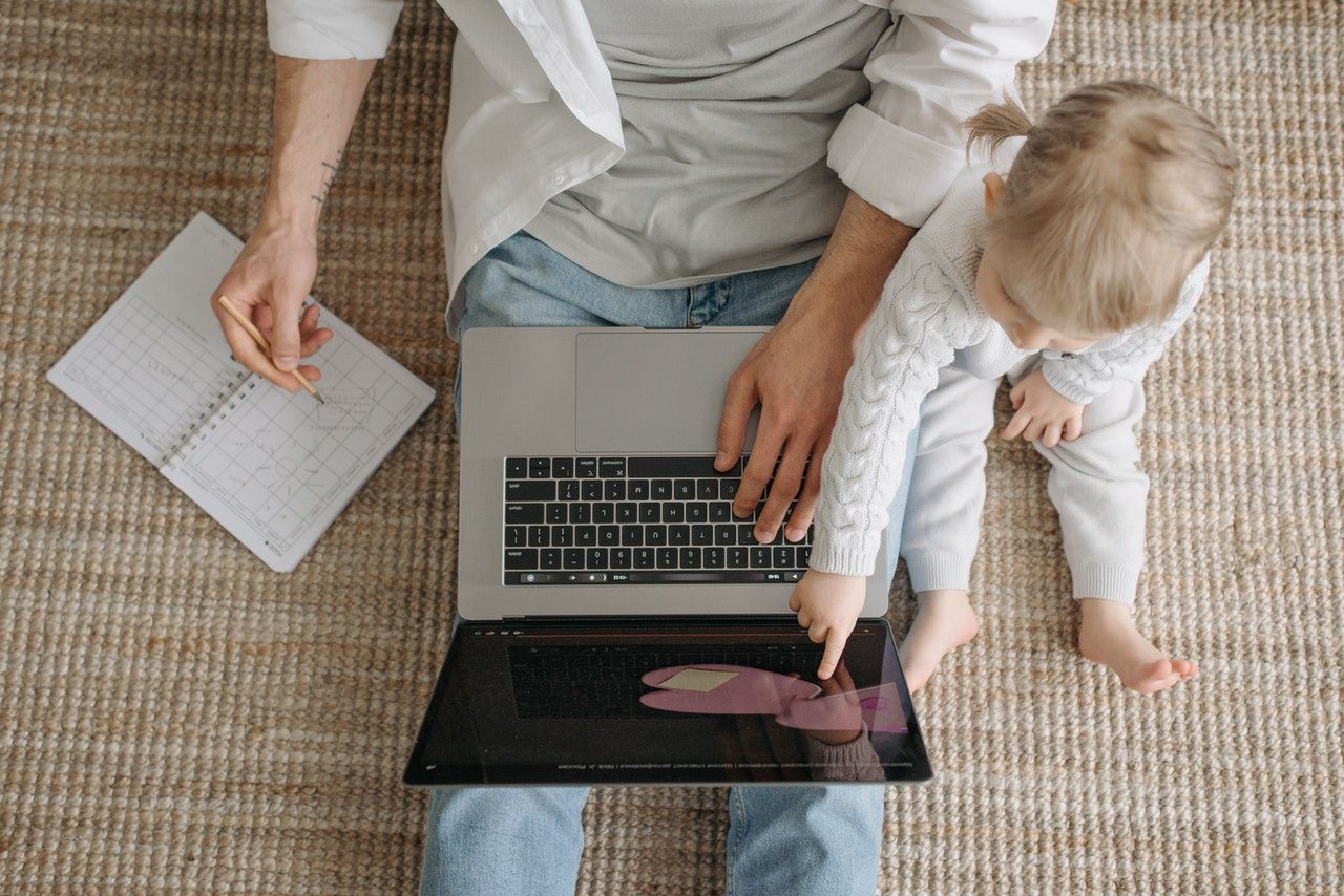 A male employee sat on the floor at home working on a laptop, with a young child in his lap. A male employee sat on the floor at home working on a laptop, with a young child in his lap.