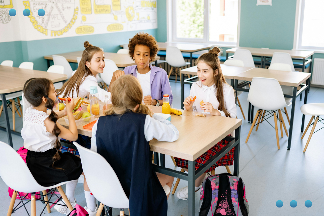 A diverse group of five students wearing school uniforms sitting around a table in a bright, modern school canteen. They are eating snacks, including fruit and sandwiches, and socializing with one another in a cheerful, well-lit environment.