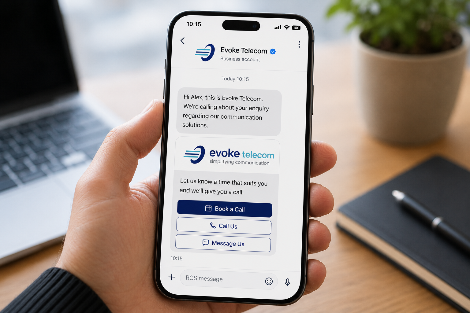 A hand holding a smartphone that displays an example of an Evoke Telecom branded RCS business message with interactive call‑to‑action buttons on a wooden desk background.