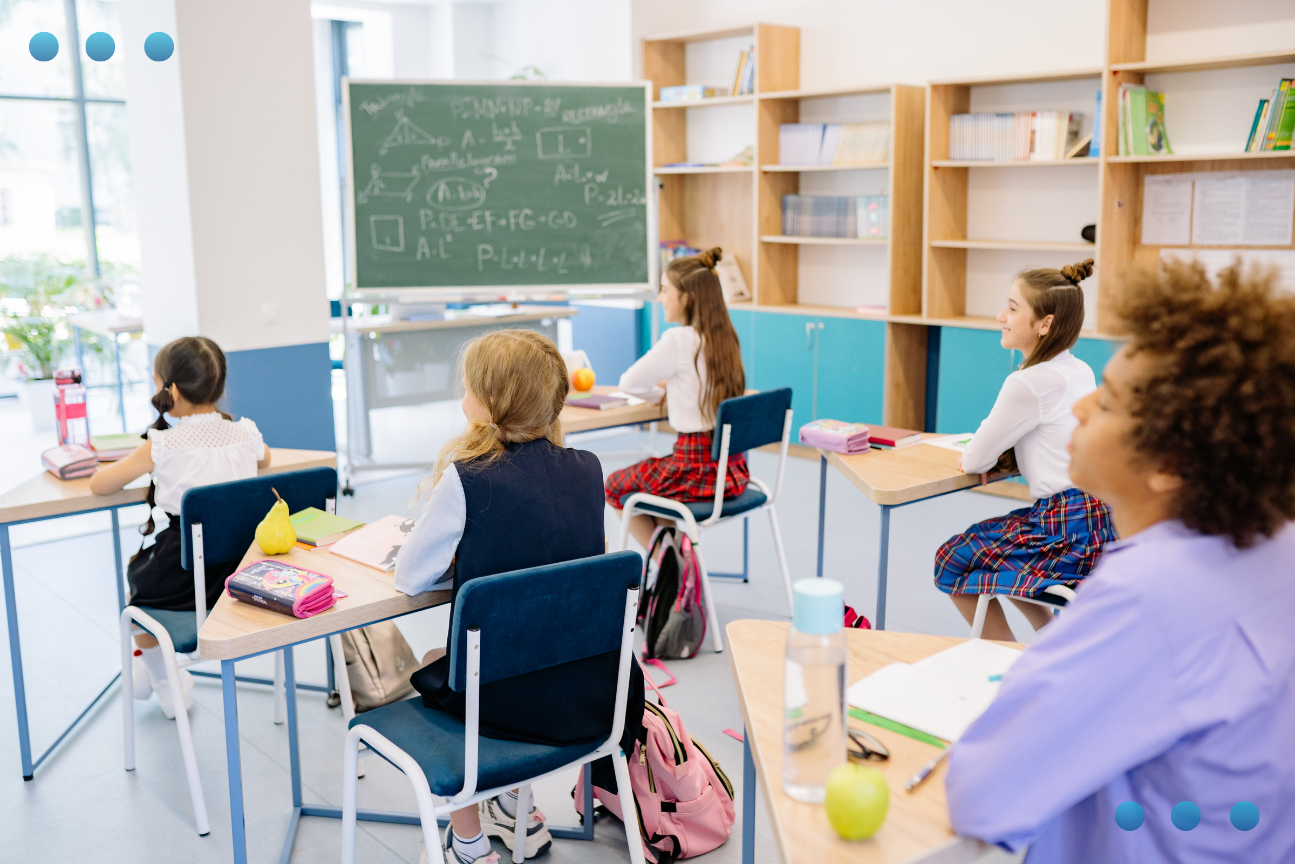 A brightly lit classroom with students seated at individual triangular desks, all facing a green chalkboard at the front covered in handwritten notes. Books, stationery, and fruit rest on the desks, and bookshelves line the back wall.