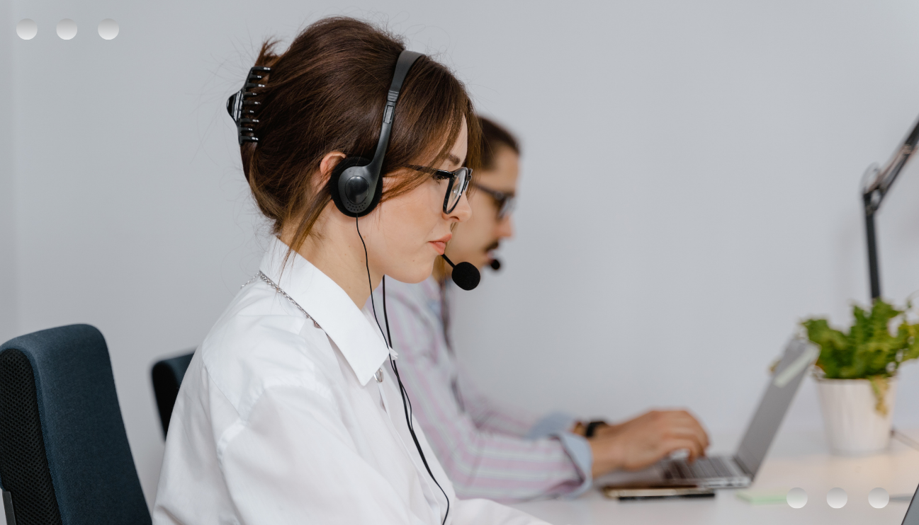 A professional contact center environment featuring a woman in the foreground wearing a headset, glasses, and a white shirt, working at a computer. In the background, a colleague is also wearing a headset and typing on a laptop