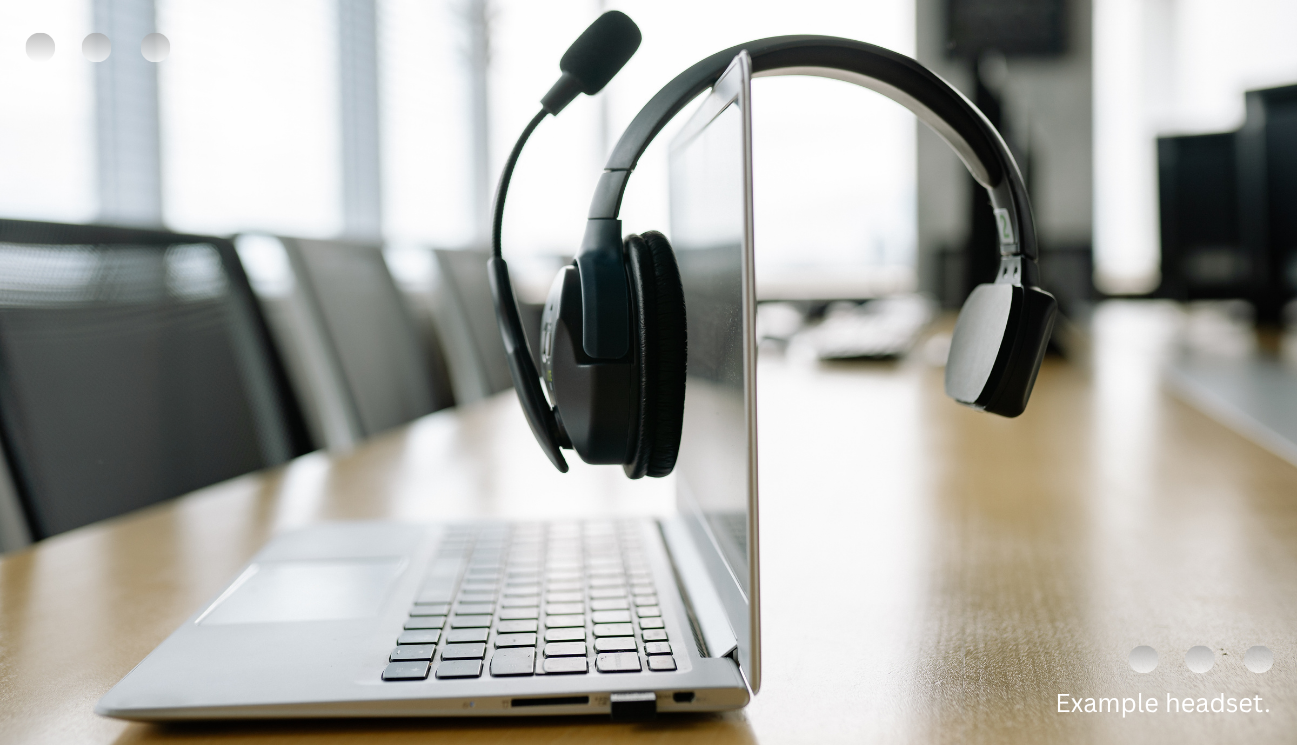 A black headset with a boom microphone draped over the screen of an open laptop, sitting on a wooden table in an office boardroom.