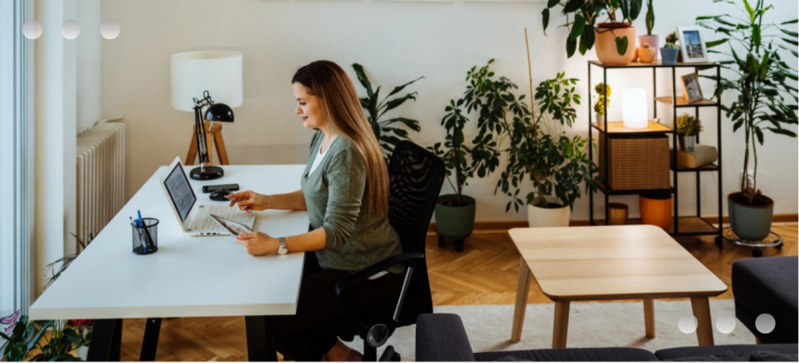 A woman working from home at a white desk, using a laptop and holding a tablet. The room is filled with numerous indoor plants, creating a bright and comfortable home office environmen