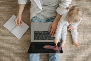 A male employee sat on the floor at home working on a laptop, with a young child in his lap.