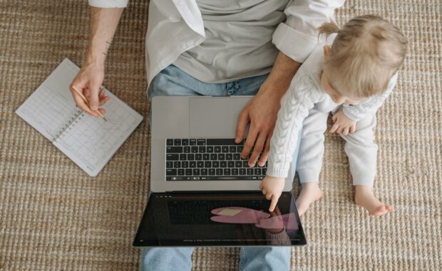 A male employee sat on the floor at home working on a laptop, with a young child in his lap.